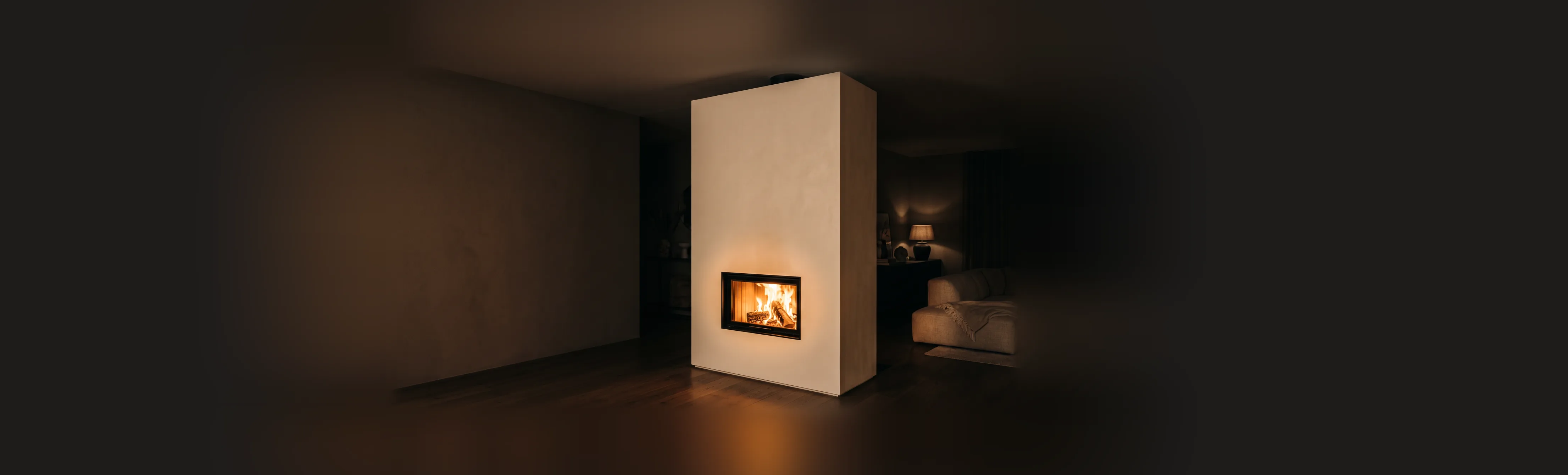 Poêle de masse contemporain, un bloc massif enduit à la chaux dans un séjour sombre du soir, sa petite porte vitrée montrant un feu vif qui éclaire l’enduit